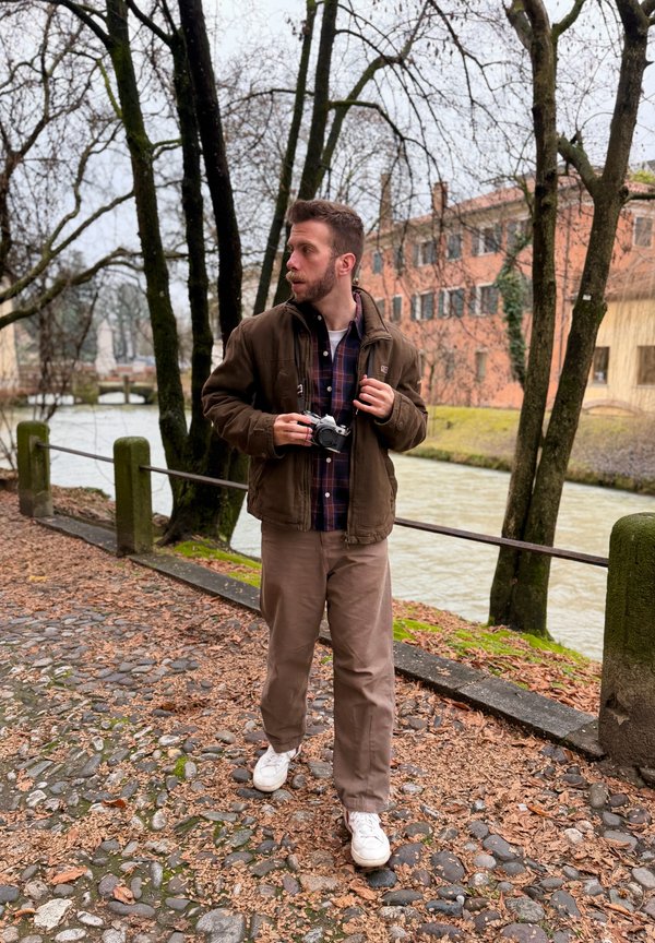 Hombre sosteniendo una cámara caminando por un camino empedrado cubierto de hojas junto a un río, con árboles desnudos y edificios al fondo.