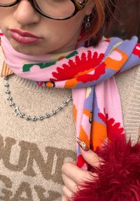 Κοντινό πλάνο ατόμου που φοράει γυαλιά με φλοράλ μοτίβο, κόκκινο κραγιόν, πολύχρωμο φλοράλ κασκόλ, μπεζ πουλόβερ, ασημένιο κολιέ με αστέρι και κόκκινο χνουδωτό μανίκι.