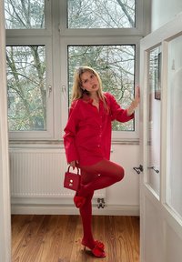 Chemise rouge surdimensionnée, collants assortis et talons avec des accents en volants. Petit sac à main rouge. Sol en bois et grandes fenêtres en arrière-plan.