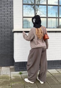 Femme portant un pull beige duveteux, un pantalon large marron, des baskets blanches, un bonnet noir avec un logo blanc, et un petit sac à bandoulière marron, debout près d'un mur en briques blanches.