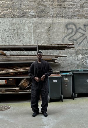Chaqueta negra de gran tamaño sobre una camisa negra, pantalones cargo y zapatos negros. Los accesorios incluyen gafas de sol y collares en capas. El fondo presenta madera apilada.