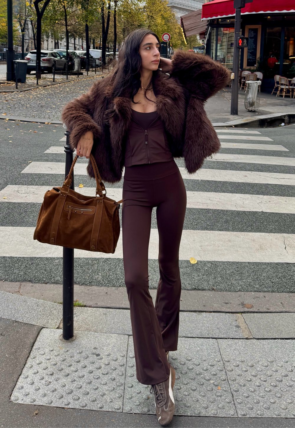 Braune Kunstpelzjacke, passendes Schokoladenoberteil und passende Hose. Braune Wildlederhandtasche und taupefarbene Schuhe mit Schnürdetails, posierend auf einem Zebrastreifen.