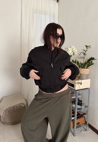 Personne portant une veste noire zippée et des lunettes de soleil, posant à l'intérieur près de plantes et d'une petite étagère en métal avec des livres et des objets décoratifs.