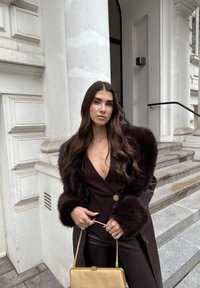 Smeđi lažni krzneni kaput sa širokim rukavima preko bluze s dubokim V izrezom, u kombinaciji s crnim hlačama i zlatnom torbicom, stoji na kamenim stepenicama.