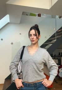 Jeune femme aux cheveux foncés attachés en chignon, portant un pull gris et un jean bleu, debout avec la main sur la hanche dans un appartement moderne avec un escalier en arrière-plan.