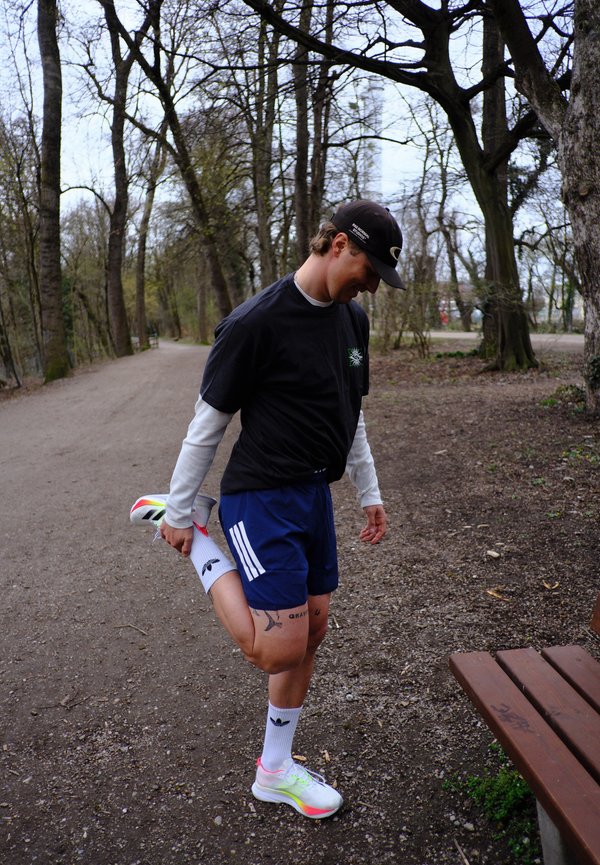 Άνδρας τεντώνει τον τετρακέφαλο τραβώντας το πόδι του πίσω σε ένα μονοπάτι στο δάσος, φορώντας αθλητικά ρούχα και μαύρο καπέλο.