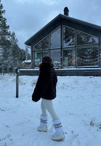 Personne en vêtements d'hiver et bottes blanches marchant sur la neige vers une maison moderne noire avec de grandes fenêtres et des guirlandes lumineuses.