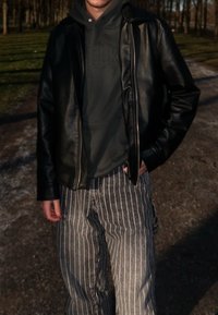 Crna kožna jakna preko tamnosive dukserice. Prugaste tamnosive traperice labavog kroja. Vanjska scena s drvećem u pozadini.