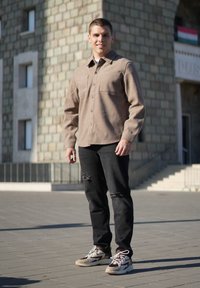 Barna gombos ing két mellzsebbel, fekete koptatott farmernadrág, és színes, vastag talpú sportcipők, kő háttér előtt.