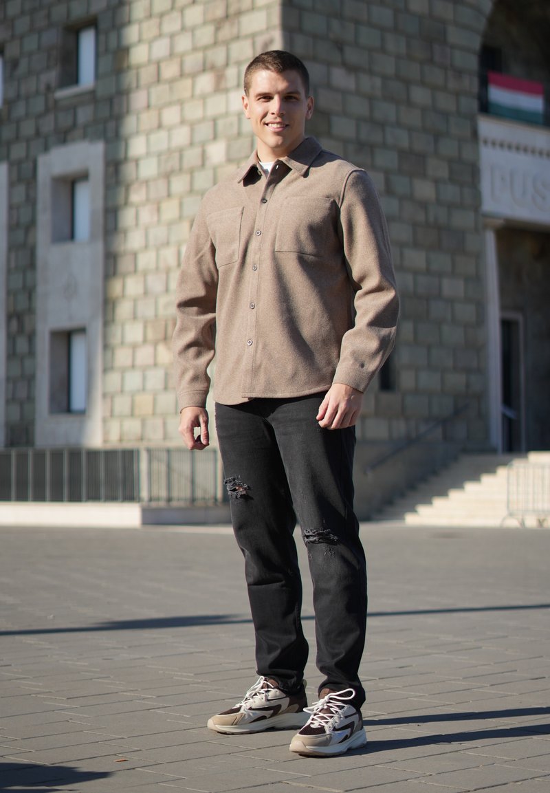 Brun skjorte med knapper og to brystlommer, sorte slidte jeans og multicolor sneakers med en kraftig sål, sat på en sten baggrund.
