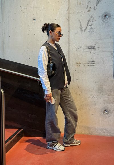 Figura femenina con un chaleco de rayas sobre una camisa blanca, jeans grises y zapatillas con estampado, sosteniendo un smartphone, cerca de una pared de concreto.