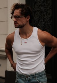 Top sin mangas de algodón blanco con logotipo rojo, combinado con pantalones de mezclilla azul claro. La persona tiene el cabello rizado y lleva accesorios en tono dorado.