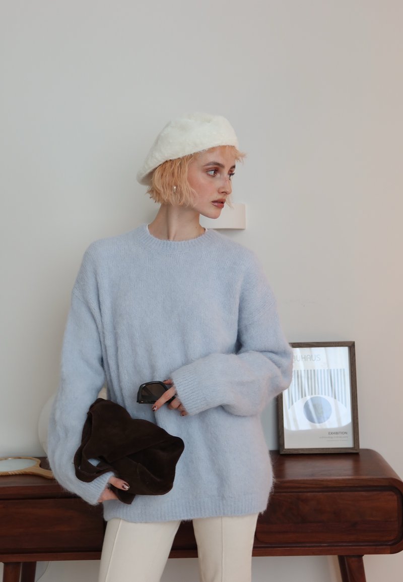 Jeune femme portant un béret blanc et un pull bleu clair, tenant un sac foncé et des lunettes de soleil, debout près d'une console en bois avec des œuvres encadrées.