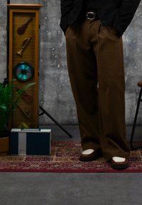 Personne portant un pantalon large marron et des chaussures marron-blancs se tenant à côté d'un haut-parleur vintage, d'une vitrine encadrée d'instruments de musique et d'une plante.