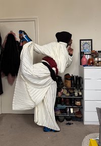 Personne en robe blanche plissée et coiffe noire, étirant la jambe en arrière, en équilibre sur un pied dans une pièce avec des chaussures et une commode.