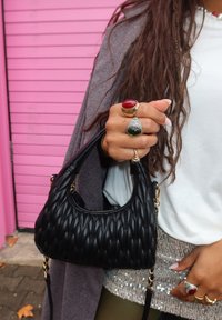 Μαύρη επενδεδυμένη τσάντα χεριού με καμπυλωτό σχήμα, χρυσά αξεσουάρ, κρατημένη από ένα χέρι διακοσμημένο με δαχτυλίδι με κόκκινο και πράσινο πολύτιμο λίθο, σε ροζ φόντο.