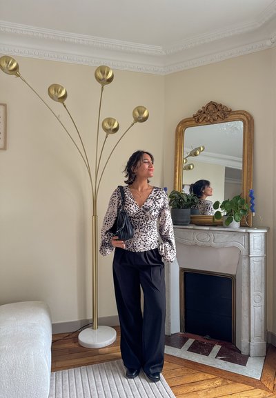 Blusa de manga larga estampada con diseño cruzado y mangas abullonadas, combinada con pantalones negros de pierna ancha. Bolso negro y lámpara de pie dorada al fondo.