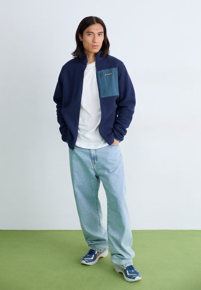 Chaqueta de forro polar en color marino con un bolsillo en el pecho de color verde azulado, combinada con jeans sueltos azul claro y zapatillas azules, contra un fondo claro.