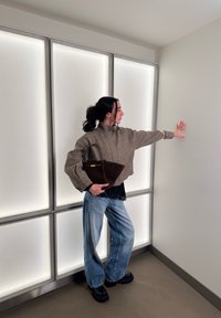 Γυναίκα με σκούρα μαλλιά σε κότσο, ντυμένη με καρό σακάκι και τζιν, κρατώντας καφέ τσάντα χειρός, αγγίζοντας έναν λευκό τοίχο σε ένα φωτεινό μοντέρνο δωμάτιο.