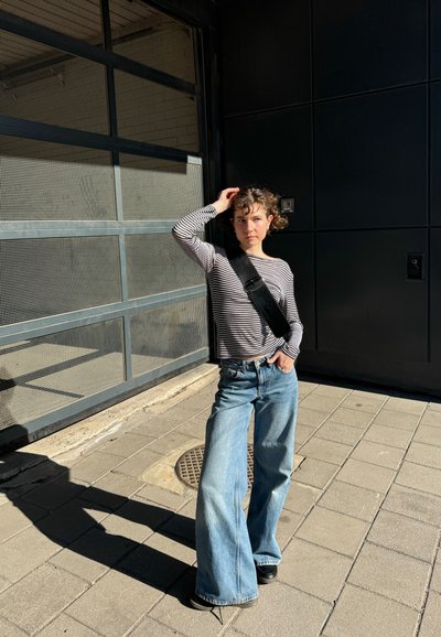 Camisa de rayas de manga larga, jeans de pierna ancha en azul claro y un bolso negro de hombro. La luz del sol proyecta una sombra sobre el pavimento.