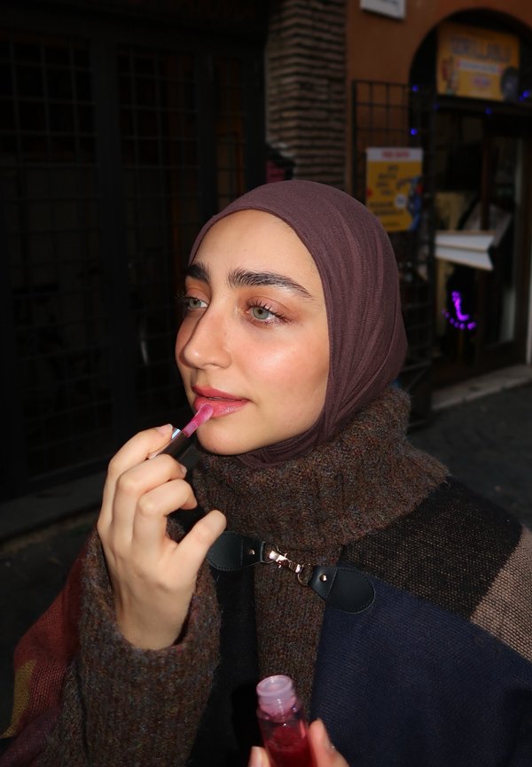 Un prodotto per labbra rosa lucido viene applicato con un applicatore trasparente. La persona indossa un hijab marrone e un maglione multicolore con texture.