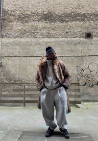 Manteau en fausse fourrure aux tons bruns, sweat à capuche gris avec fermeture éclair, pantalon de survêtement gris et baskets noires, posés devant un mur texturé gris.