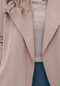 Femme portant un manteau en laine beige sur un pull beige et un pantalon foncé, avec des cheveux blonds lâches visibles au col.
