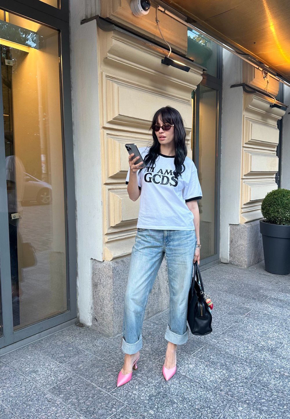 Λευκό T-shirt με κοντά μανίκια και μαύρο κείμενο "I AM GCDS", ανοιχτό μπλε τζιν με ρολό, ροζ μυτερές γόβες, μαύρη τσάντα, που στέκεται στο πεζοδρόμιο.