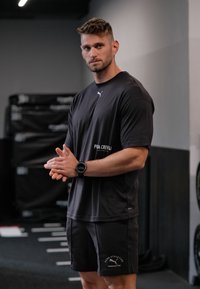 Czarna sportowa koszulka i szorty wykonane z tkaniny, z logo. Osoba nosi czarny smartwatch i stoi z założonymi rękami.