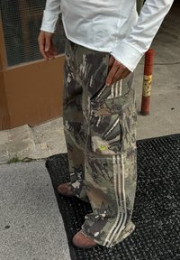 Camo Cargo-Hosen in Grüntönen und Grau, ausgestattet mit mehreren Taschen und drei weißen Streifen an der Seite, sowie einem kleinen grünen Logo.