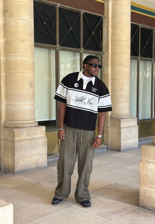 Homme portant des lunettes de soleil, tee-shirt noir et blanc "All In", pantalon cargo olive, debout entre des colonnes en pierre dans un couloir carrelé.