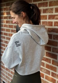 Sweat à capuche gris avec une coupe décontractée et une capuche ; présente un texte noir « WORLD SERIES OF FITNESS RACING » au dos. Porté avec des leggings foncés.