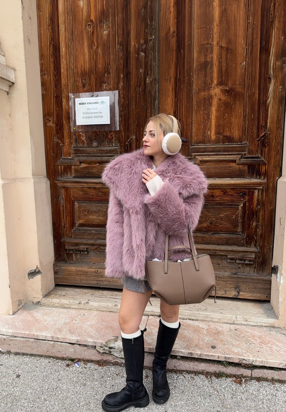 Femme portant un manteau en fausse fourrure rose, des cache-oreilles beige, des bottes montantes noires et une jupe grise, se tenant devant une grande porte en bois, tenant un sac à main taupe.