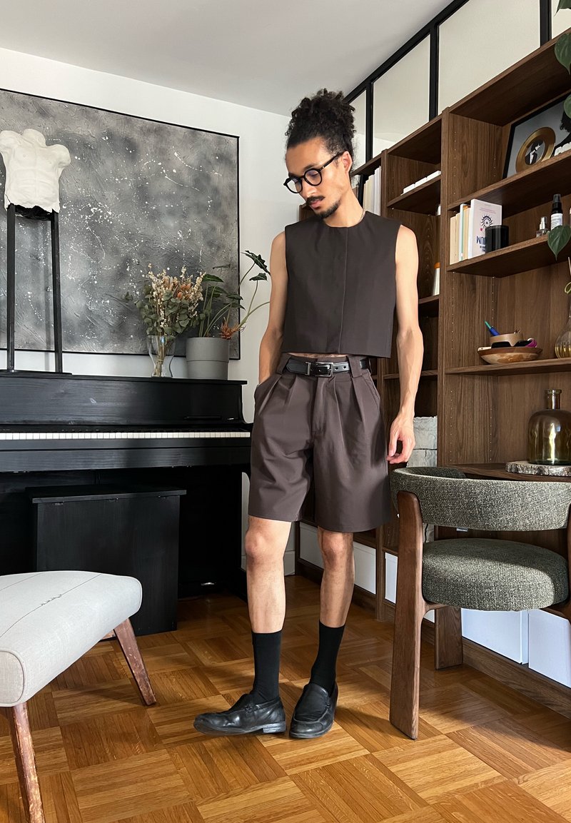 Homme avec lunettes, portant un haut noir sans manches et un short, debout près d'un piano noir et d'une bibliothèque en bois dans un salon moderne.