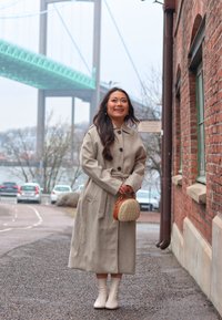 Light gray wool coat with buttons and a belt, worn over beige ankle boots. Small round wicker handbag, brick wall, and bridge in background.