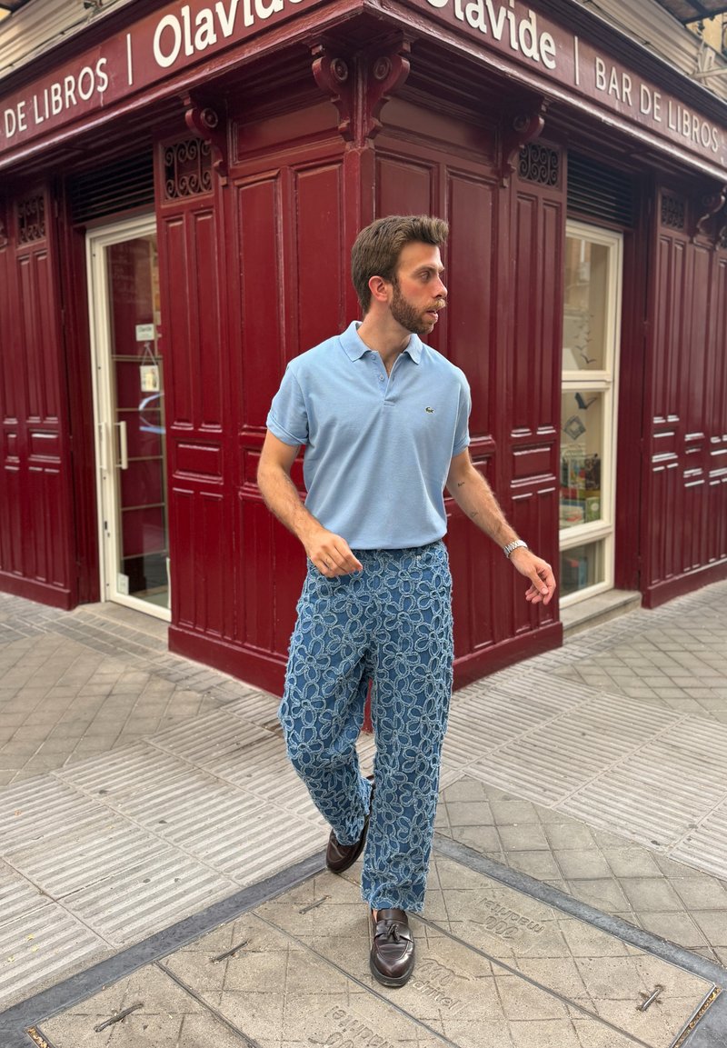 Homme en polo bleu et pantalon à motifs marchant devant un bâtiment d'angle rouge portant l'étiquette "Olavide Bar de Libros" dans une rue pavée.