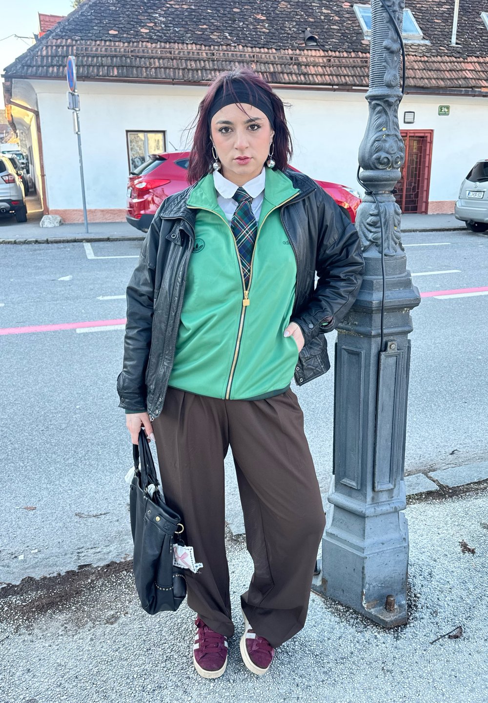 Giacca verde con zip sopra una camicia bianca e cravatta a quadri, abbinata a pantaloni larghi marroni e sneakers bordeaux. Borsa nera.