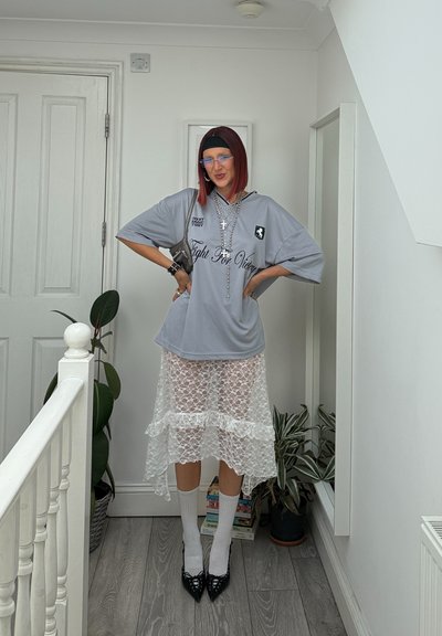 Camisa gris de gran tamaño con texto, superpuesta a una falda de encaje blanco transparente. Tacones negros altos, calcetas blancas hasta la rodilla y collares plateados en capas.