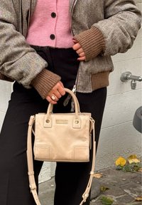 Personne portant une veste marron et un pull rose, tenant un sac à main beige avec un pantalon noir, debout près d'un mur avec un robinet.