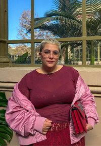 Giacca bomber in satin rosa sopra un top bordeaux, abbinata a una gonna coordinata. Il soggetto tiene una borsa a mano rossa con hardware decorativo.