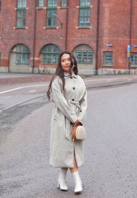 Light gray, long wool coat with tie waist and buttons, paired with white ankle boots and a small woven handbag. Smooth texture, tailored fit.