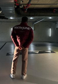 Μαρέν φουσκωτό μπουφάν με λευκό λογότυπο "SIXTHJUNE", μπεζ παντελόνια και λευκά sneakers. Αποτυπωμένο σε ένα καλά φωτισμένο, βιομηχανικό χώρο.