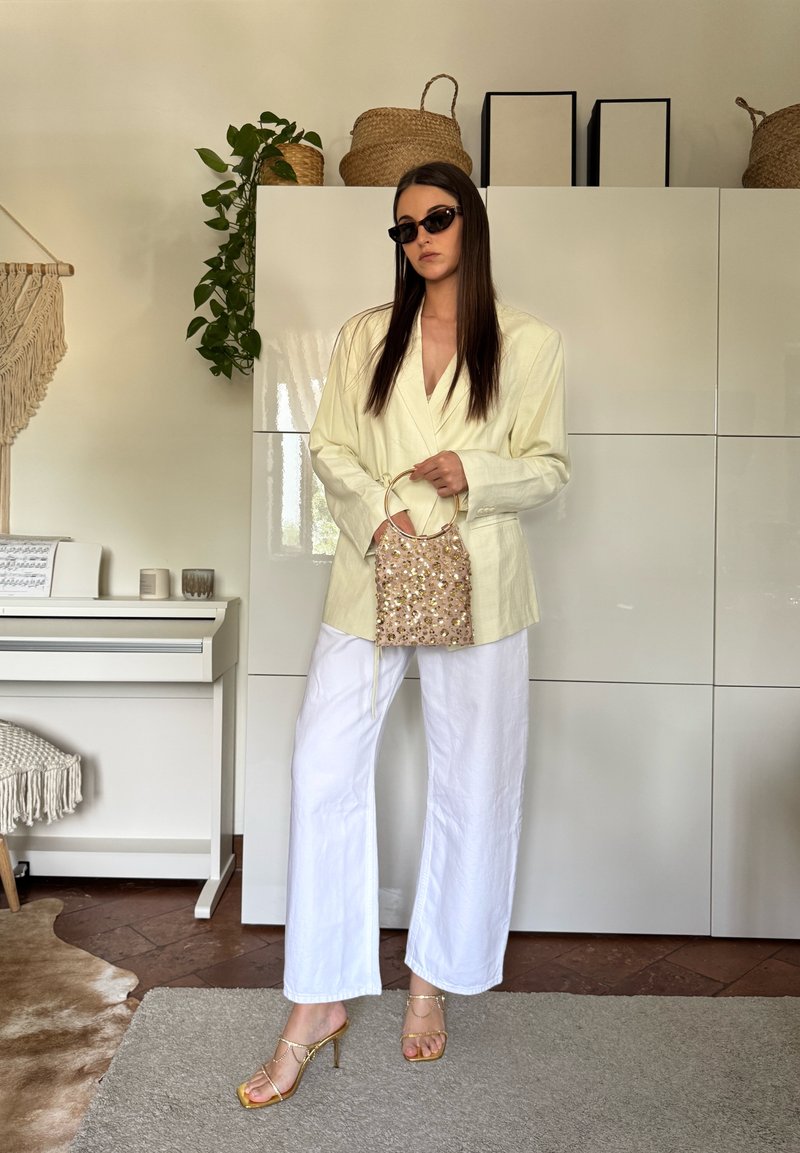 Femme portant un blazer jaune pâle, un pantalon blanc à jambes larges, des talons dorés et des lunettes de soleil, tenant un sac à sequins dans un salon.