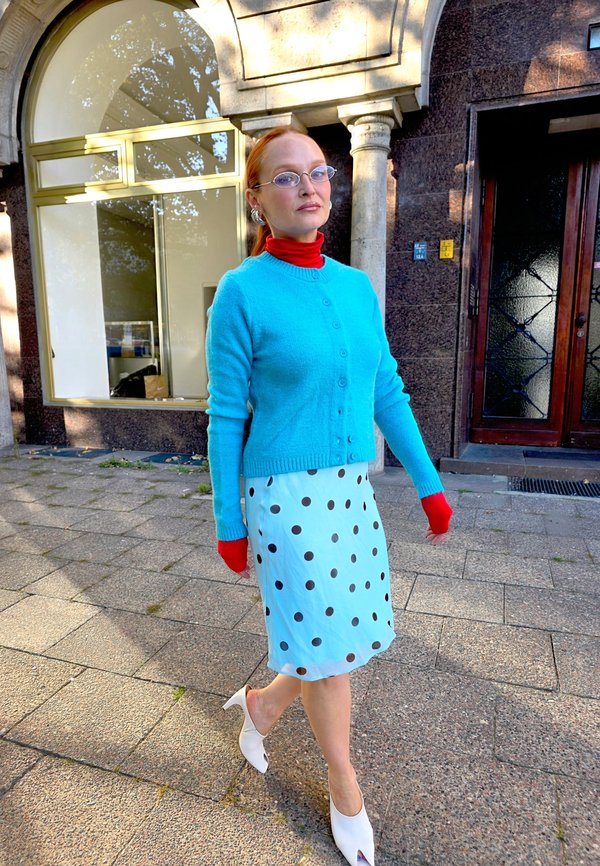 Femme aux cheveux roux portant des lunettes, un col roulé rouge, un pull bleu, une jupe à pois bleu clair et des talons blancs marchant sur un trottoir pavé près d’un bâtiment en pierre.
