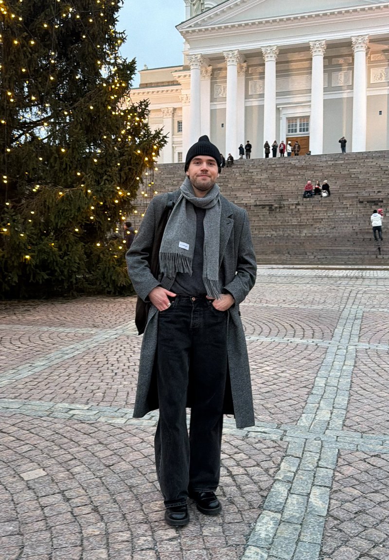 Homme portant un manteau gris, une écharpe et un bonnet noir se tenant sur une place pavée près d'un grand sapin de Noël illuminé ; bâtiment historique et escaliers en arrière-plan.