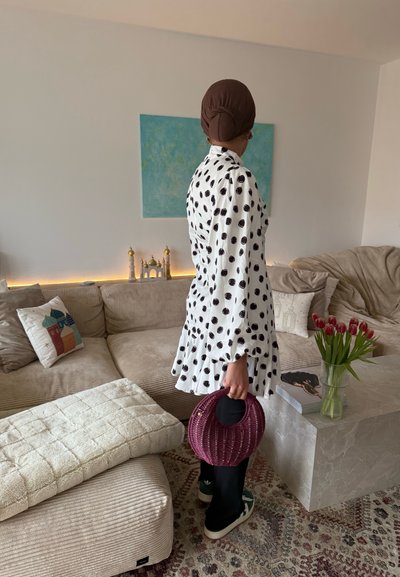 Mujer con pañuelo marrón en la cabeza y vestido blanco con lunares, sosteniendo un bolso redondo morado, de pie en una sala de estar con sofá beige y jarrón con tulipanes.