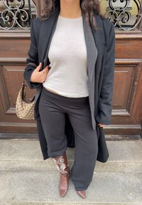 Čierny dlhý kabát, svetlosivá košeľa, čierne široké nohavice, hnedé čižmy s bielou výšivkou a vzorovaná kabelka. Drevené pozadie.