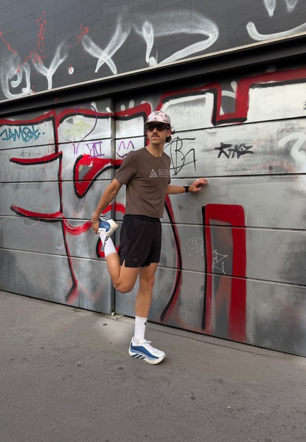 Giovane uomo in abbigliamento sportivo che allunga una gamba tenendosi a un muro metallico coperto di graffiti per supporto su un marciapiede urbano.