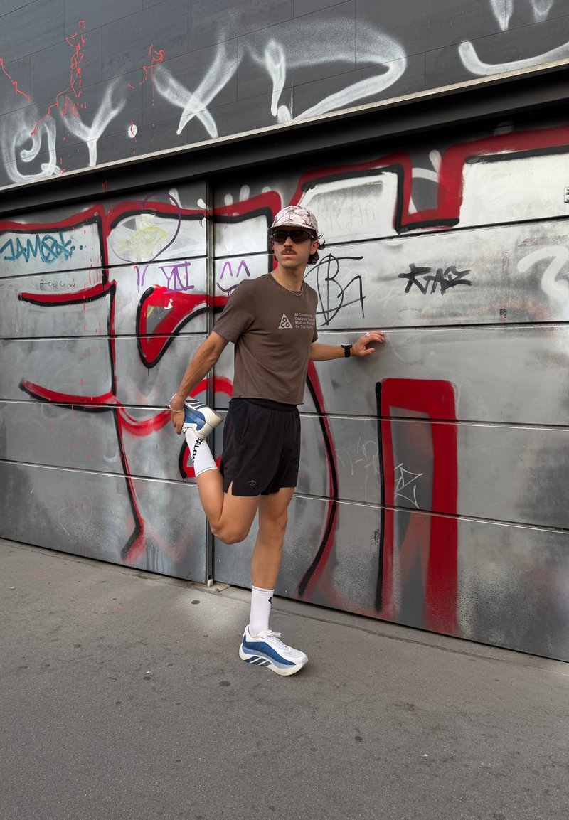 Jeune homme en tenue de sport étirant une jambe en tenant un mur métallique couvert de graffitis pour s'appuyer sur un trottoir urbain.