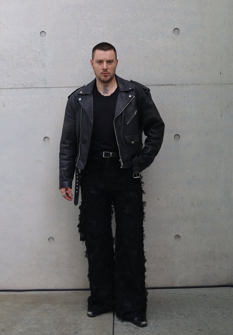 Veste de moto en cuir noir avec quincaillerie argentée, associée à un pantalon noir texturé avec des détails en franges. En arrière-plan, un mur gris.